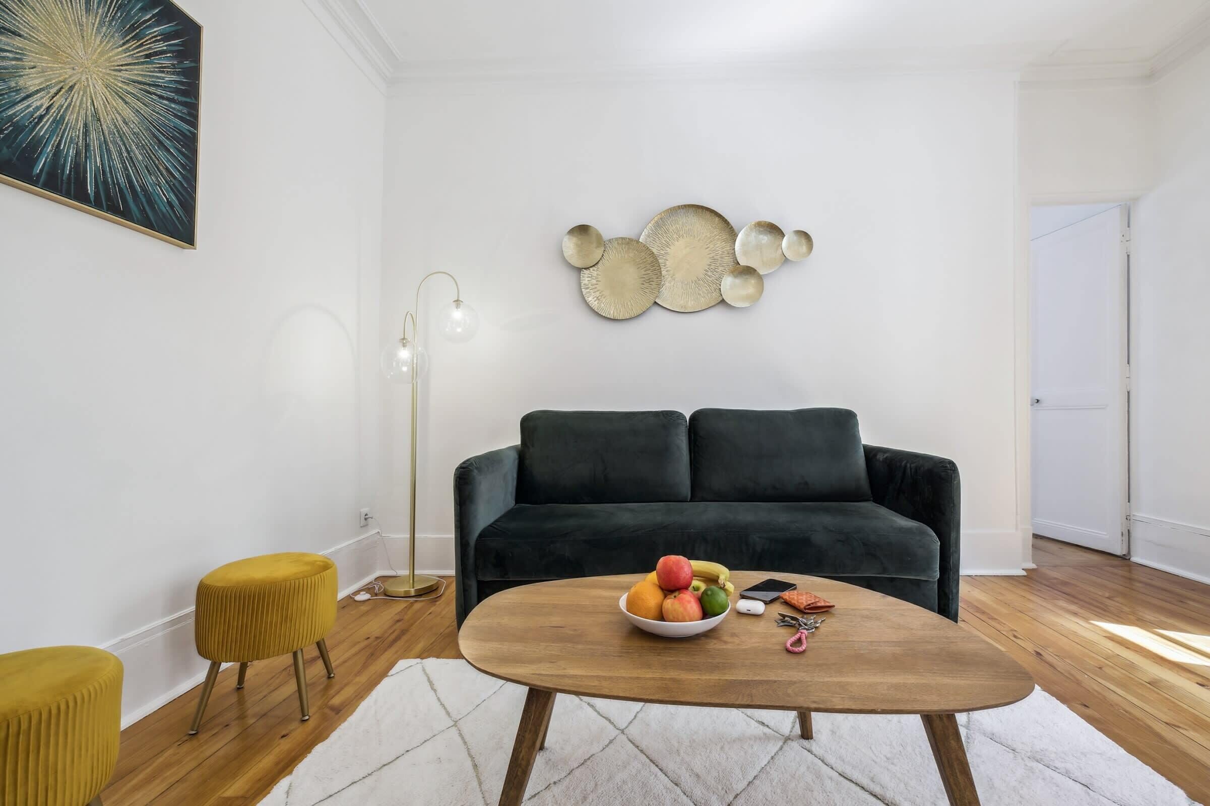 Een moderne woonkamer met een donkergroene bank, een houten salontafel met een schaal fruit, een geel voetenbankje en een wanddecoratie in abstracte stijl.