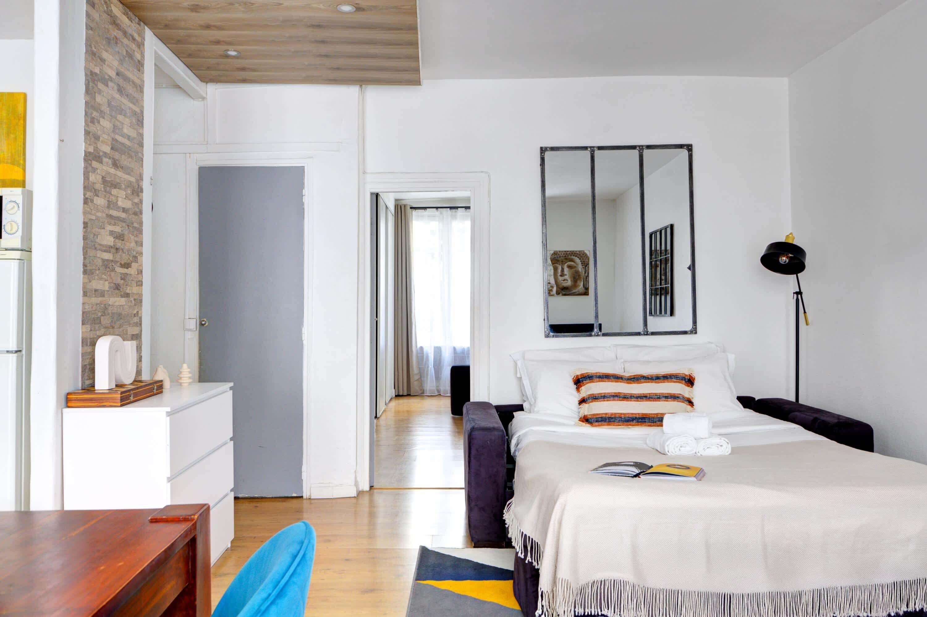 A modern hotel room with a wooden ceiling, a bed with a white and striped blanket, a bedside table with a book, a small desk with a lamp, and a mirror on the wall.