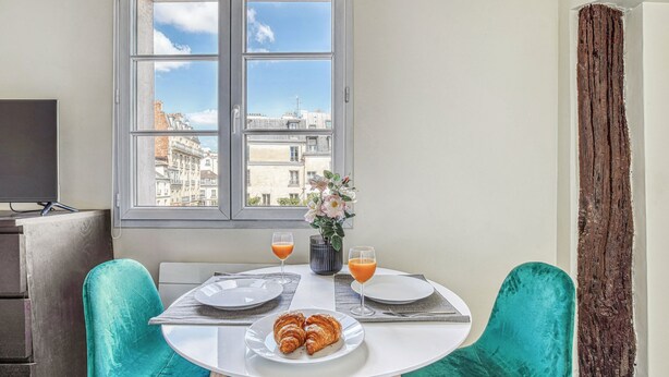 A dining area with a table set for two, a window with a view of buildings, and a vase with flowers. - Cozy Studio in The Heart of Latin Quarter (Paris)