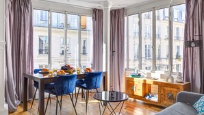 A modern room with a dining table, blue chairs, a sofa, and a small cabinet. Large windows offer views of buildings outside. - Stylish 2BR Steps from Louvre - Place Vendome (Paris)