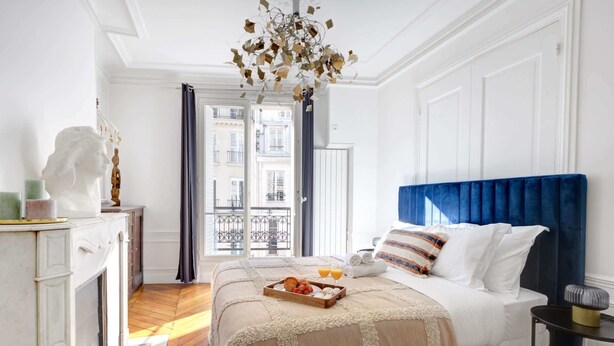 A hotel room with a bed, a side table, a fireplace, and a view of the outside. - Stylish 2BR Steps from Louvre - Place Vendome (Paris)