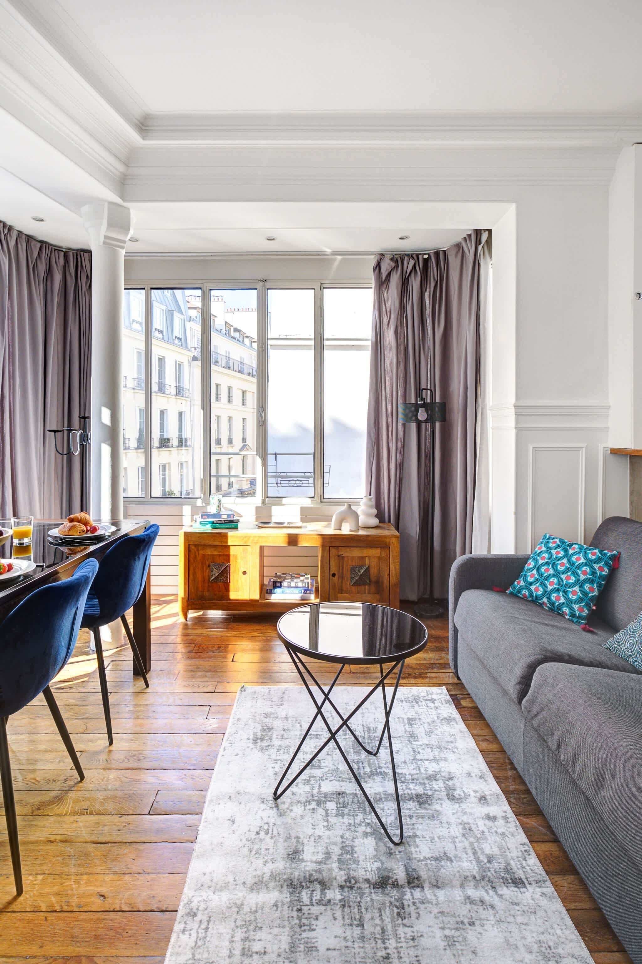 A modern living room with a sofa, a dining table with chairs, a coffee table, and a view of buildings through large windows.