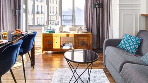 A modern living room with a sofa, a dining table with chairs, a coffee table, and a view of buildings through large windows. - Stylish 2BR Steps from Louvre - Place Vendome (Paris)