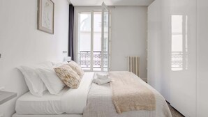 A minimalist bedroom with a bed, a radiator, and a window with a balcony view. - Stylish 2BR Steps from Louvre - Place Vendome (Paris)