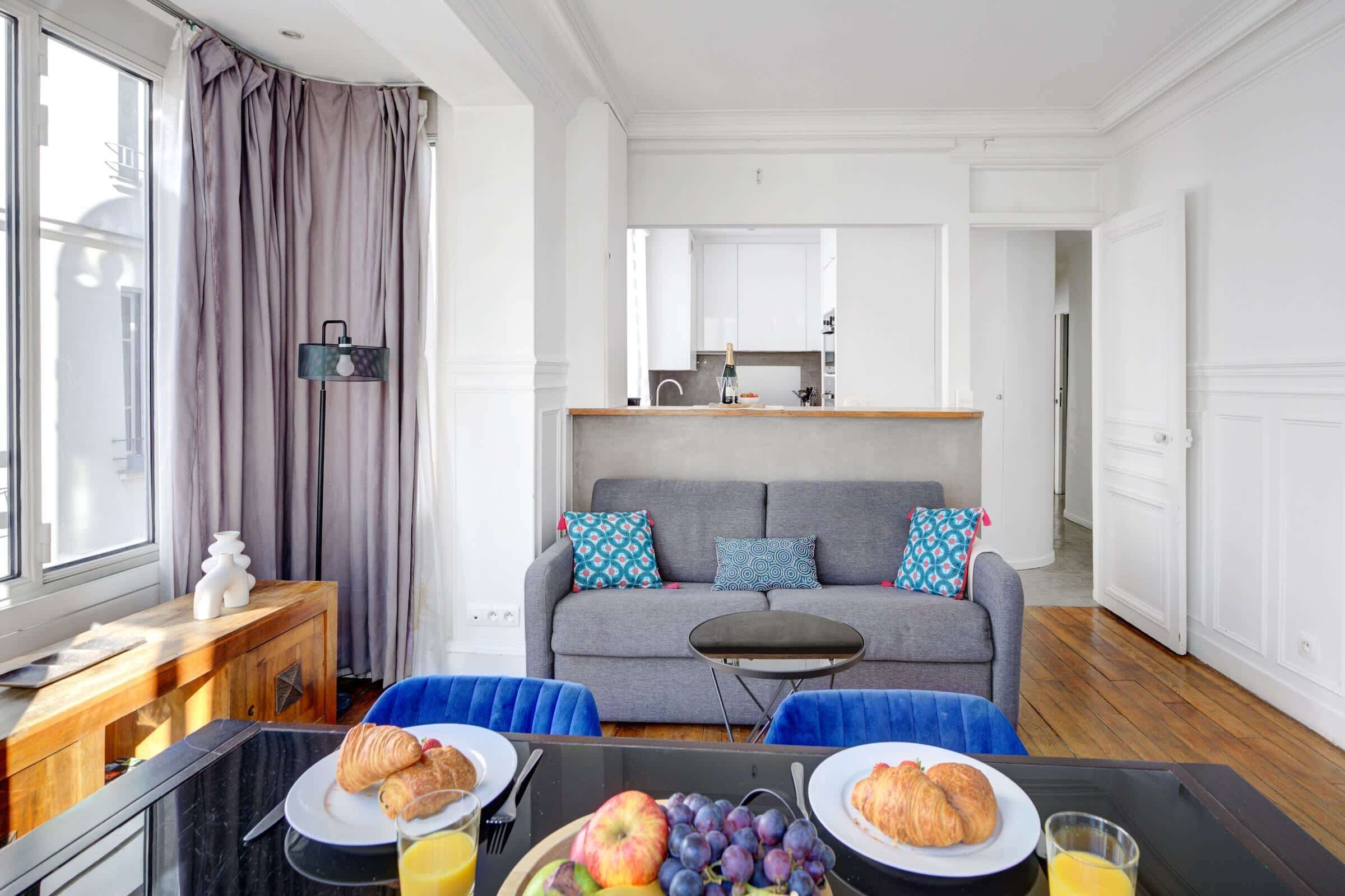 En moderne stue med en grå sofa, blå stole, et lille bord med morgenmad og et kig til køkkenet i baggrunden.