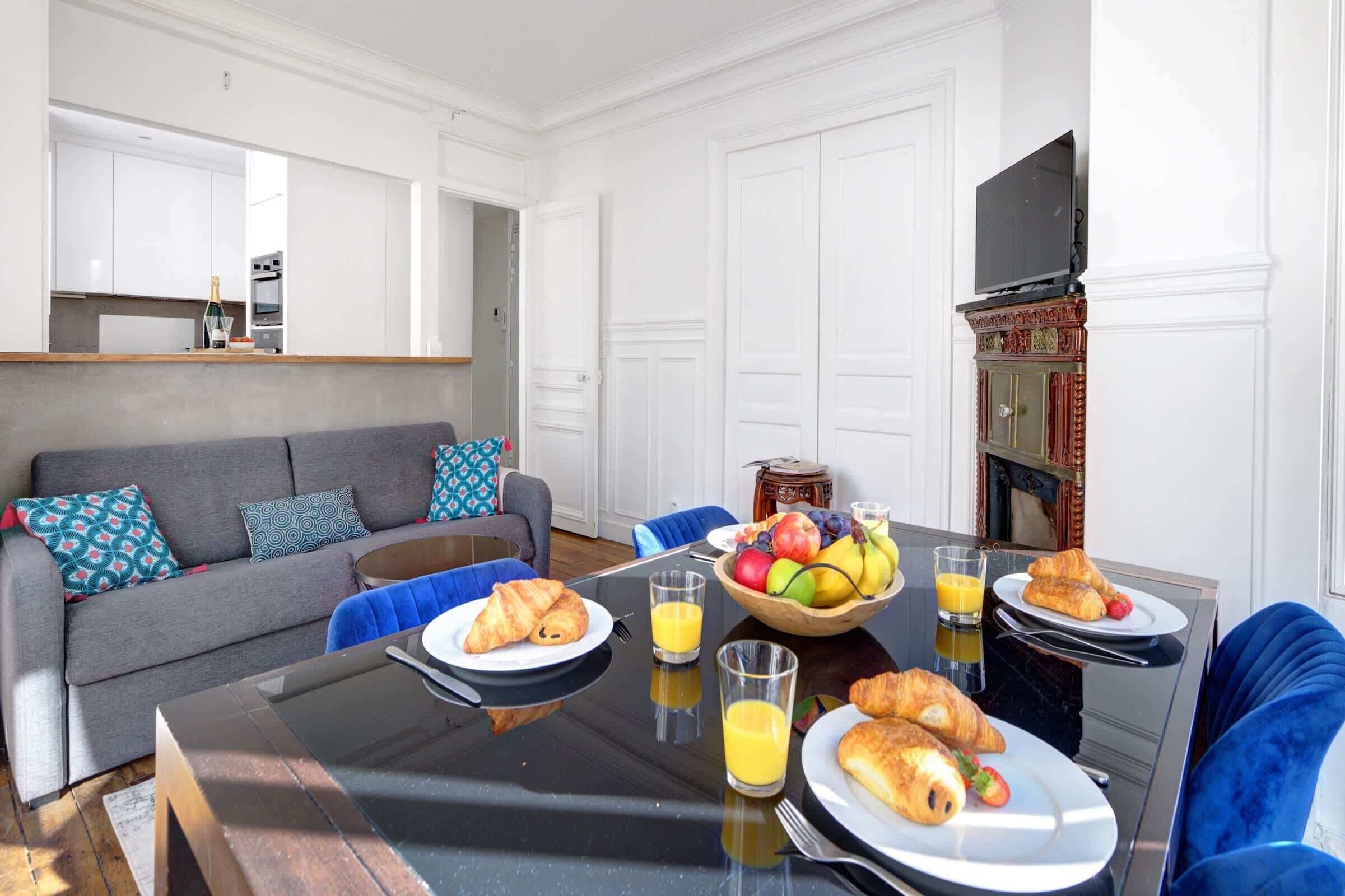 A dining area with a table set for breakfast, including croissants, fruit, and juice. There is a sofa and a television in the background.