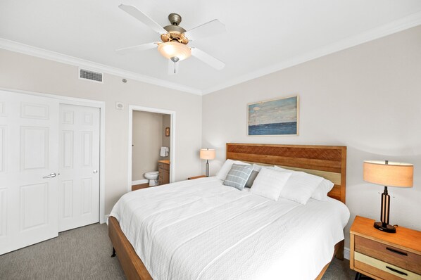 A bedroom with a bed, a ceiling fan, a nightstand, a lamp, a painting on the wall, and a door.