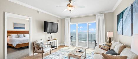 Een hotelkamer met een bed, een televisie, een balkon met uitzicht op de oceaan, een plafondventilator en een groot schilderij aan de muur.