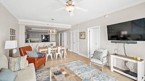 A spacious living room with a ceiling fan, a TV mounted on the wall, a dining area with a table and chairs, and a kitchen in the background. - Beachfront-Pool-Balcony-Wonderful View (Gulf Shores)