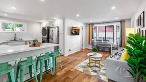 A modern kitchen with white cabinets, a central island with bar stools, a dining area with a table and chairs, and a living room with a sofa and a plant. - Hidden Renovated Garden Cottage Near Costco, I99 (Shoreline)