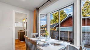 A dining area with a white table set for four, large windows, and a view of a wooden deck and trees. - Hidden Renovated Garden Cottage Near Costco, I99 (Shoreline)