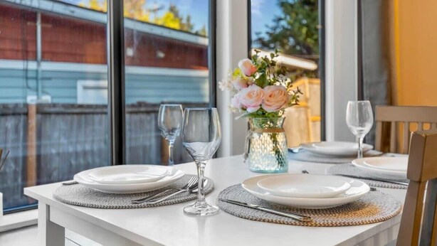 A dining table set with plates, cutlery, and wine glasses, with a vase of flowers and a view of a building outside. - Hidden Renovated Garden Cottage Near Costco, I99 (Shoreline)