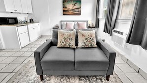 Una sala de estar moderna con un sofá gris, dos cojines decorativos, un cuadro de un atardecer y una zona de cocina con armarios blancos y un microondas.