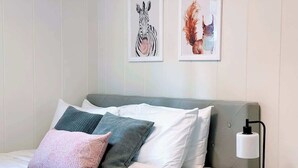 A neatly made bed with white bedding and multiple pillows, a bedside table with a lamp, and two framed artworks on the wall.