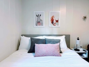 Un lit bien fait, avec une literie blanche et grise, deux tableaux représentant des animaux encadrés au mur, et une table de chevet avec une lampe et une télécommande.