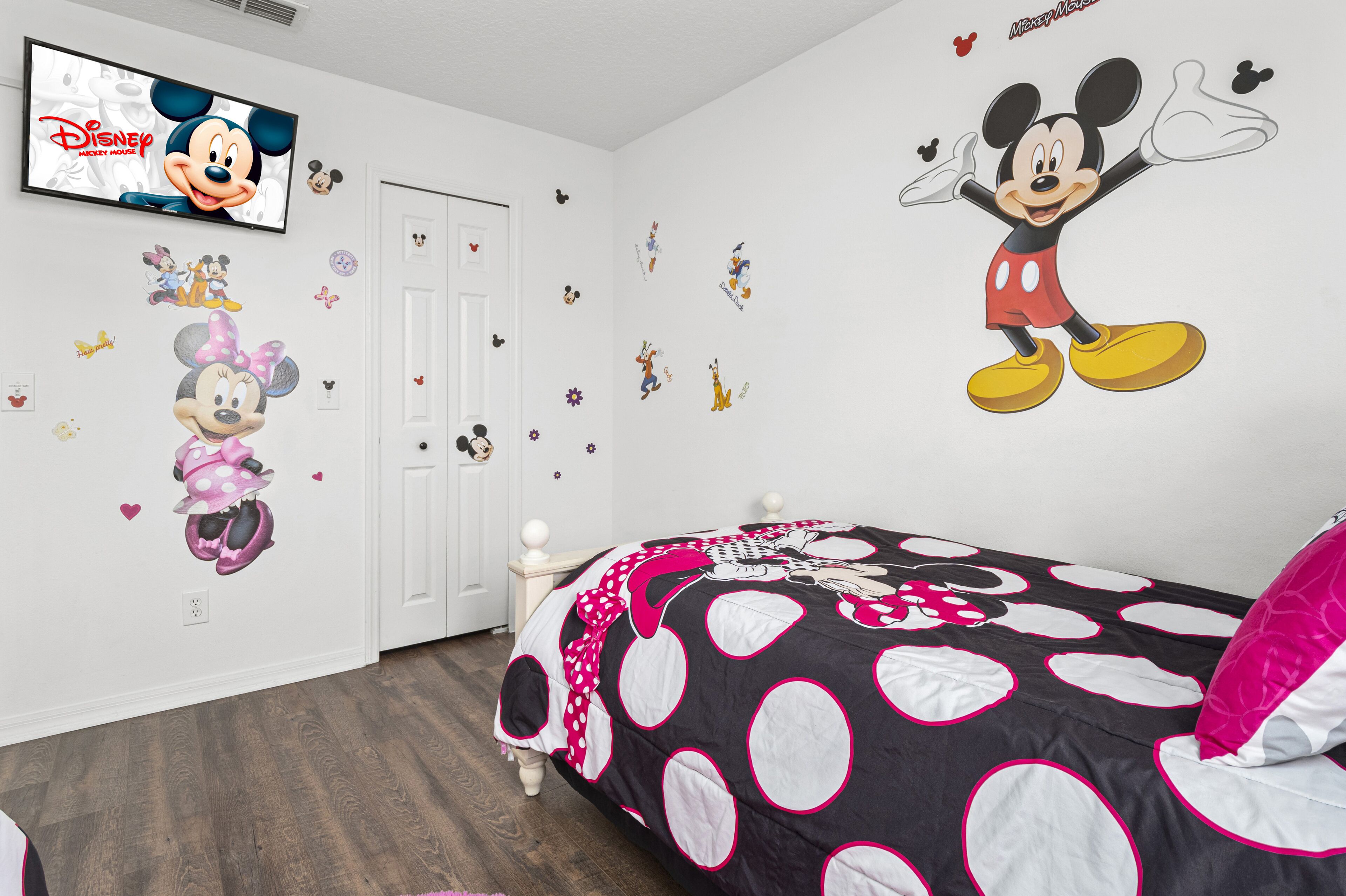 Une chambre décorée avec des autocollants de Mickey et Minnie sur les murs, un lit recouvert d’une couette à pois et aux motifs de Mickey, et un téléviseur affichant le logo Disney de Mickey Mouse.