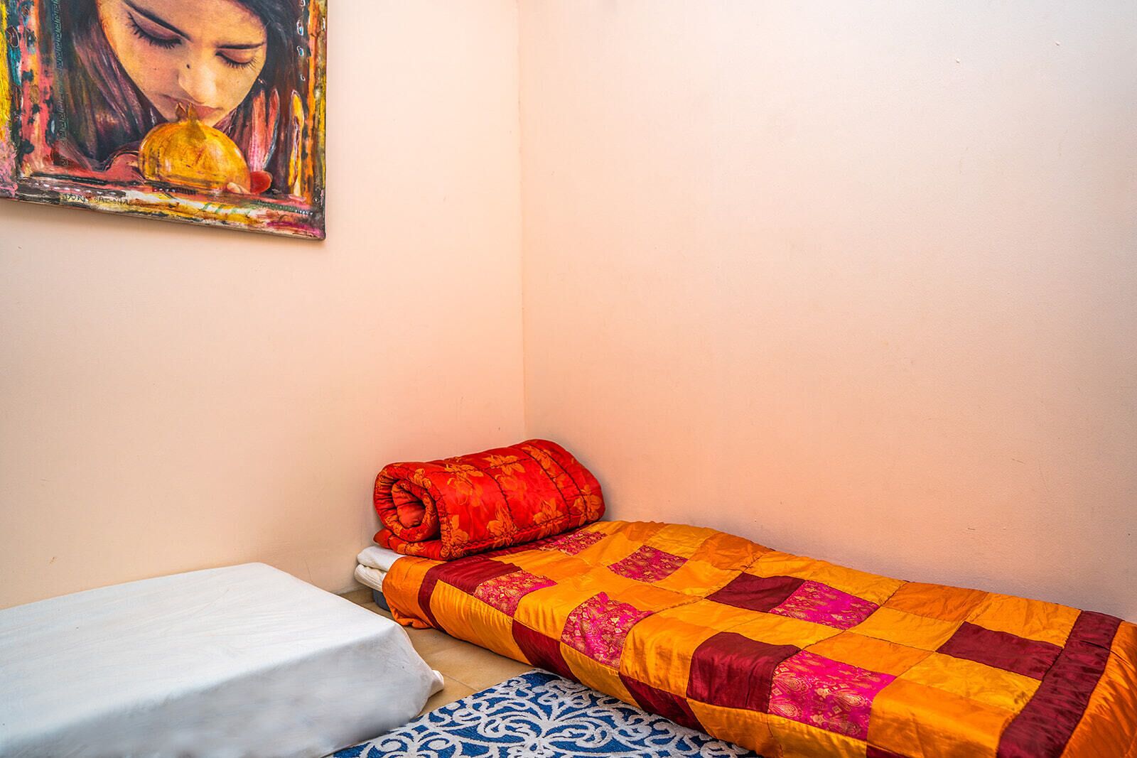 A small, neatly made bed with a red and yellow quilt, a white pillow, and a blue patterned pillow.