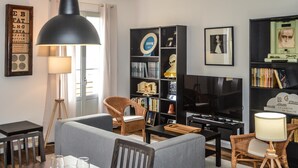 Moderni olohuone, jossa on sohva, kahdelle katettu ruokapöytä, televisio ja kirjahylly, joka on täynnä kirjoja ja koriste-esineitä.