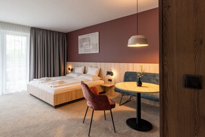 A modern hotel room with a large bed, a round table, a sofa, and a wall-mounted lamp.
