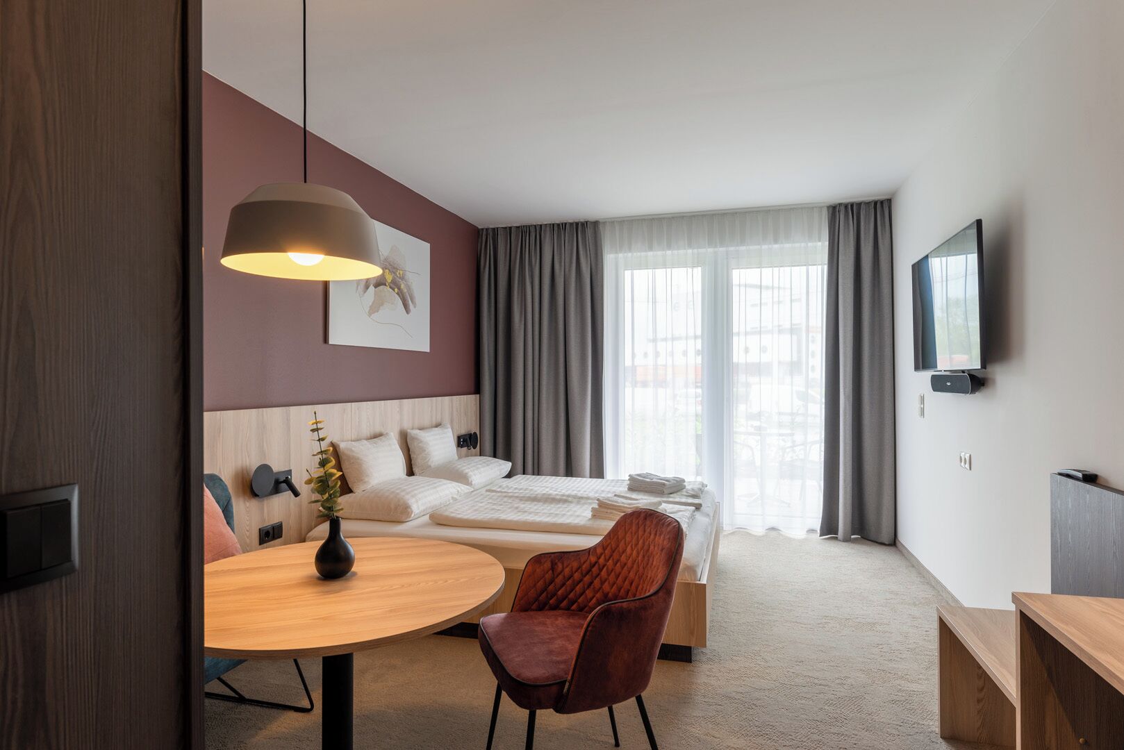 A modern hotel room with a bed, a desk, a chair, a round table, a TV, and a window with curtains.