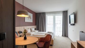 A modern hotel room with a bed, a desk, a chair, a round table, a TV, and a window with curtains.