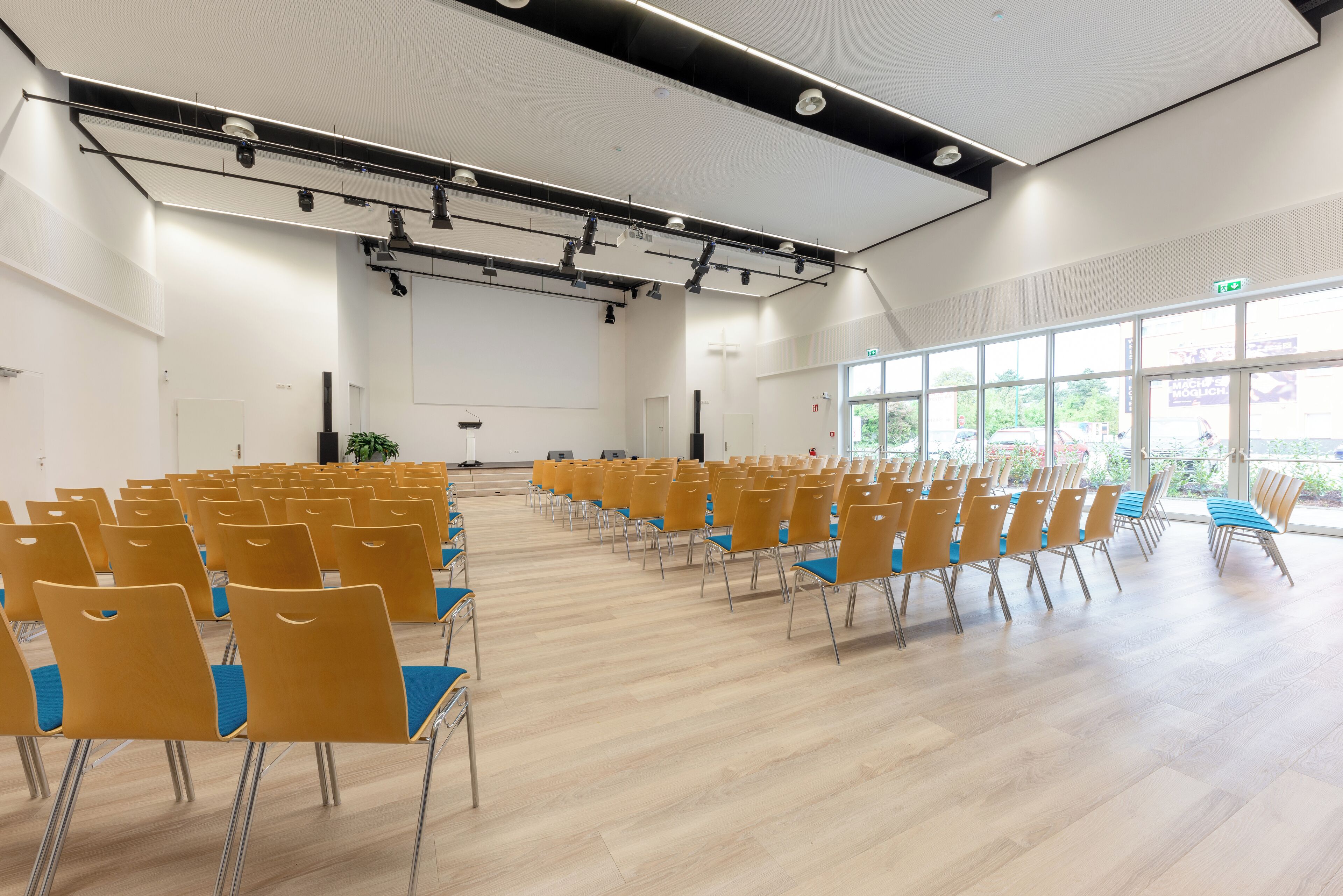 Un amplio salón con hileras de sillas, un escenario, ventanas grandes y un diseño de techo moderno.