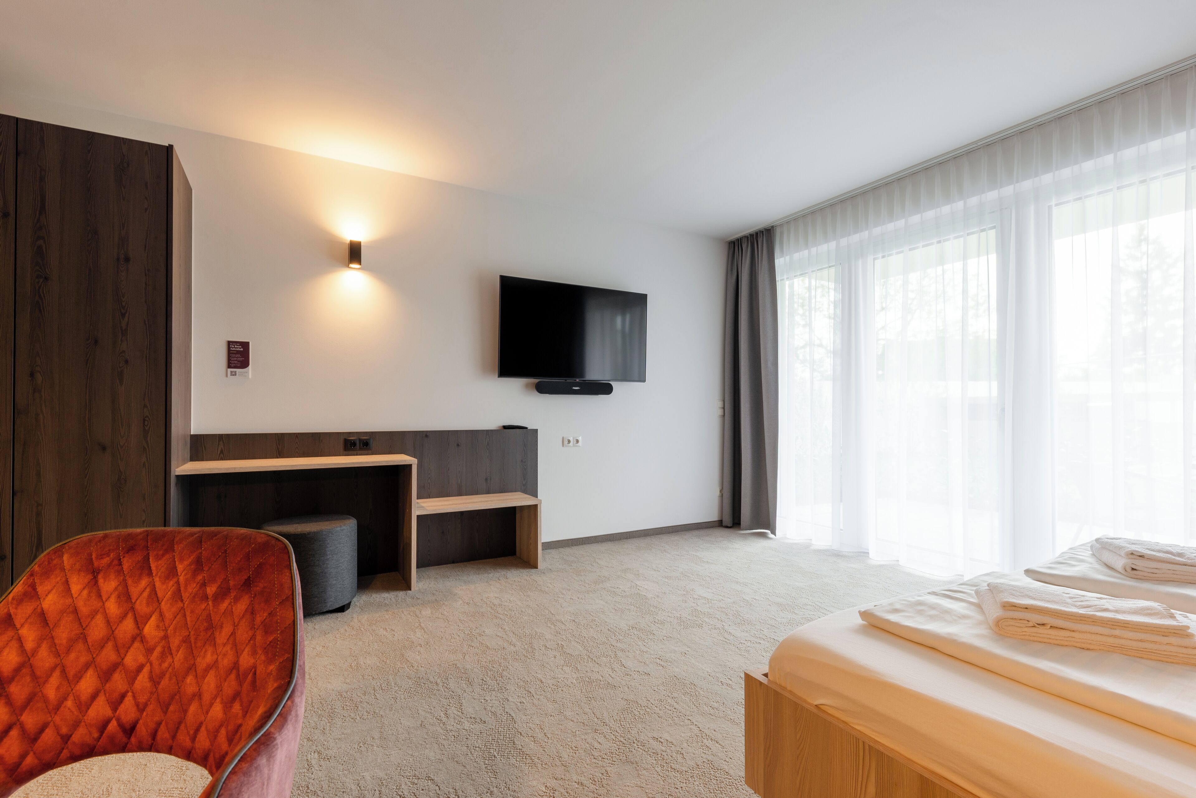 Ein Hotelzimmer mit Bett, einem Wandfernseher, einer Sitzecke mit einer Bank und einem großen Fenster mit Vorhängen.