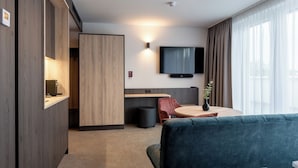 A modern hotel room with a wooden wardrobe, a flat-screen TV mounted on the wall, a round dining table with a red chair, and a blue sofa. - Hotel Novapart (Wels)
