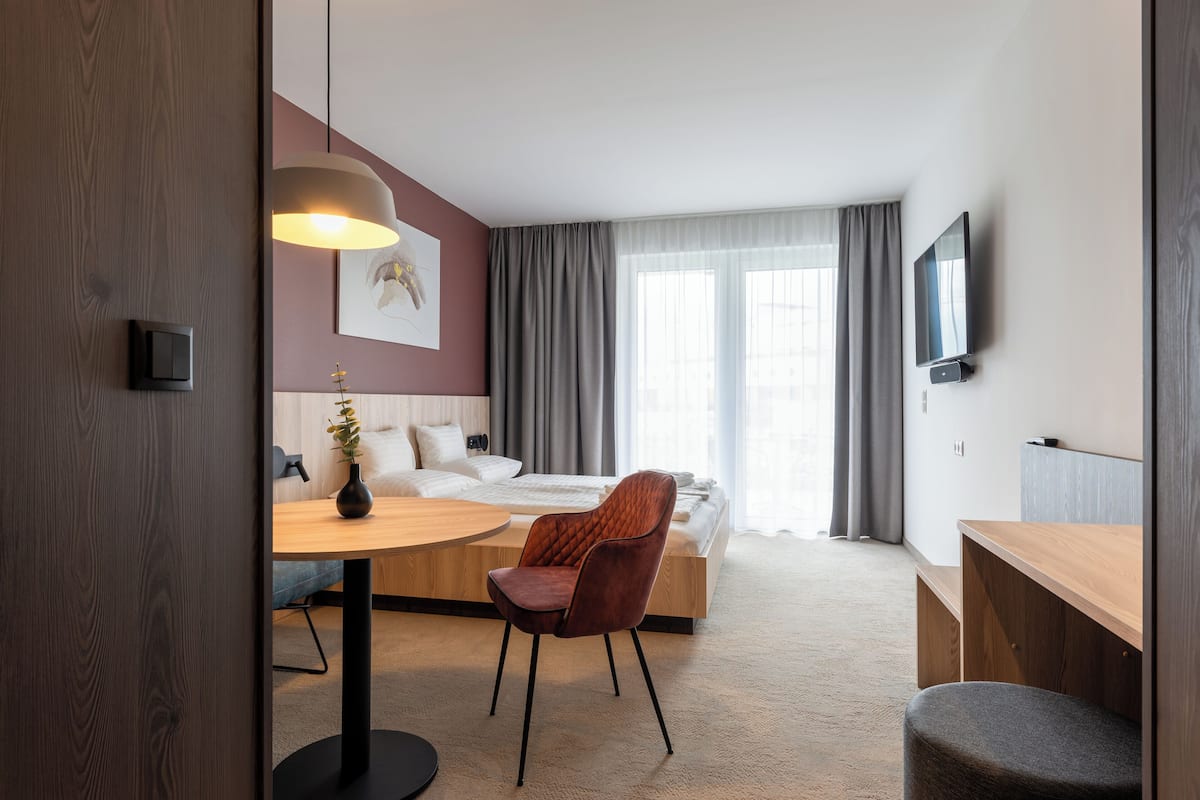 A modern hotel room with a wooden desk, a comfortable armchair, a bed with a white headboard, a TV mounted on the wall, and a large window with curtains.