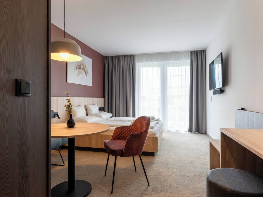 A modern hotel room with a wooden desk, a comfortable armchair, a bed with a white headboard, a TV mounted on the wall, and a large window with curtains.