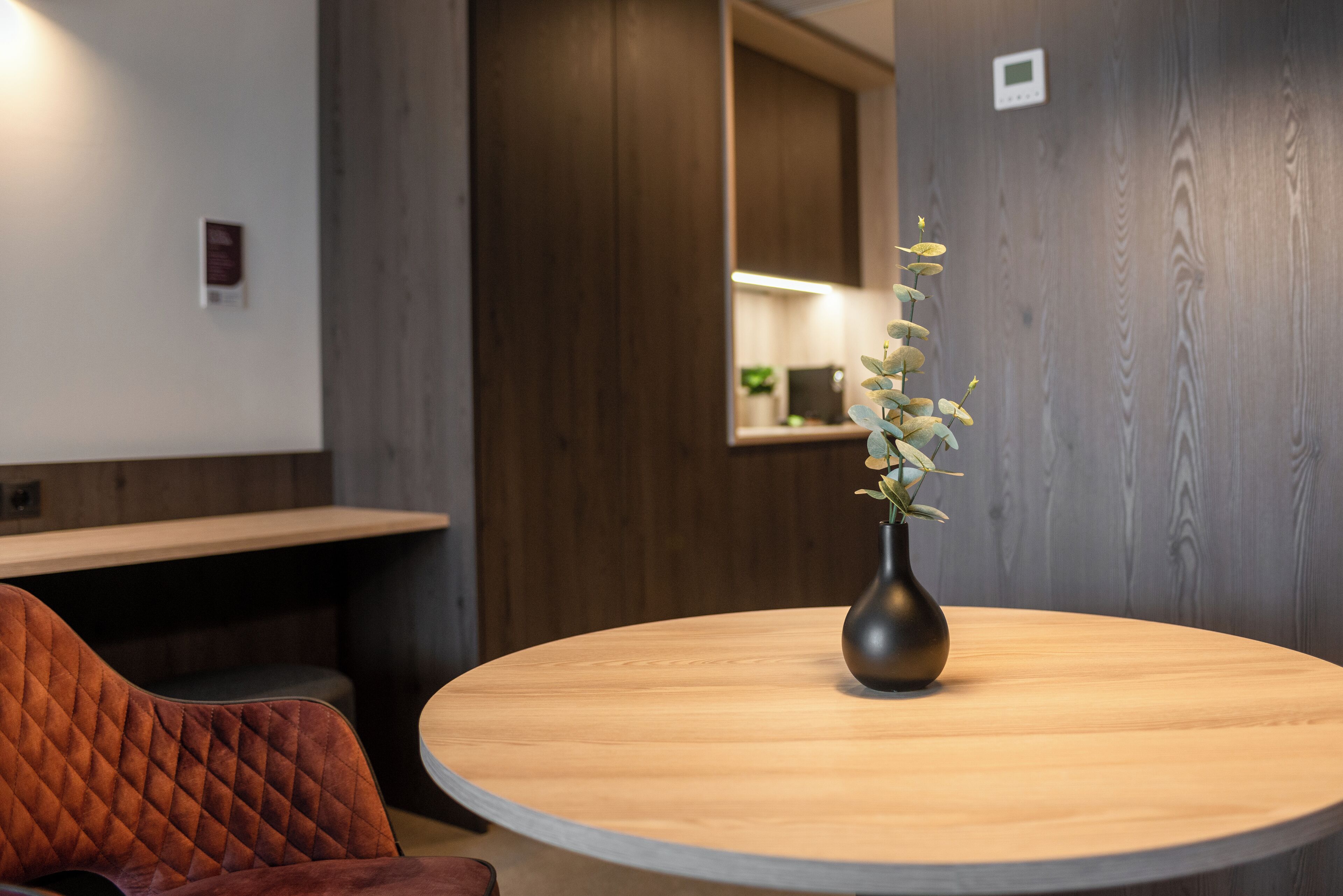 Una mesa redonda de madera con un florero oscuro que contiene flores blancas, una silla con cojín y una pared de madera con un letrero pequeño.