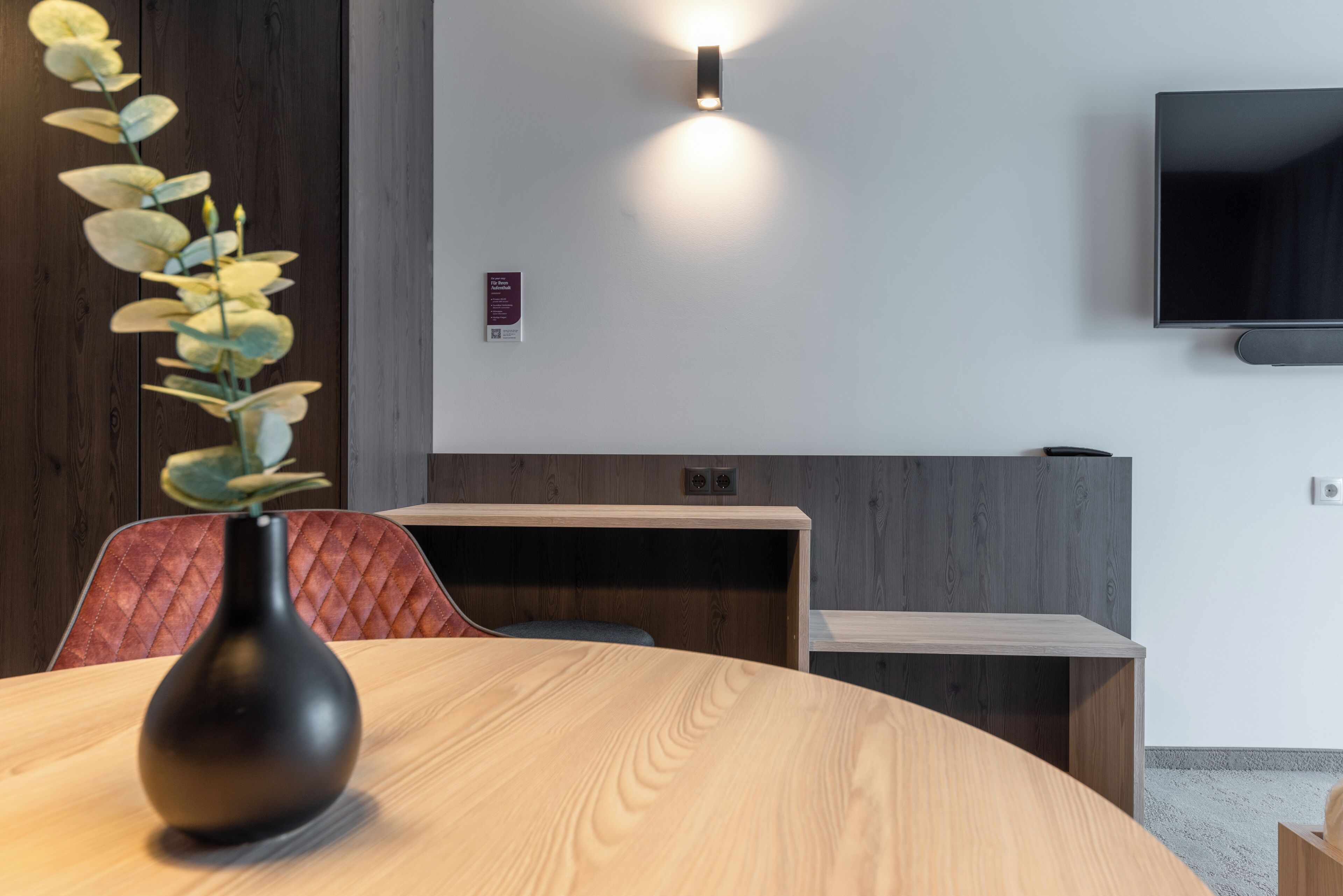 Een vergaderruimte met een ronde tafel, een zwarte vaas met bloemen, een aan de muur bevestigde televisie en een houten scheidingswand.