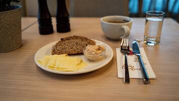 Ein Teller mit Brot, Butter, einer Aufstrichcreme, eine Tasse Kaffee, ein Glas Wasser und Besteck auf einem Holztisch.
