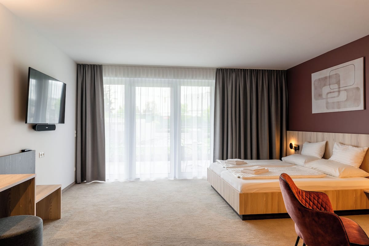 A modern hotel room with a large bed, a flat-screen TV mounted on the wall, a desk with a chair, and a view of the outdoors through large windows.