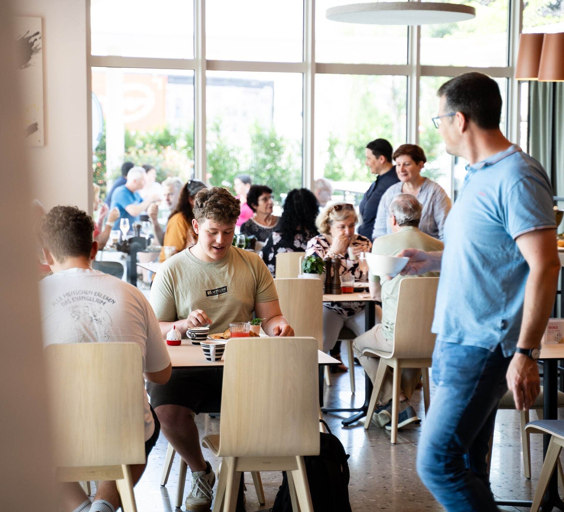 Een informeel restaurant met meerdere tafels en stoelen, grote ramen en een licht interieur.