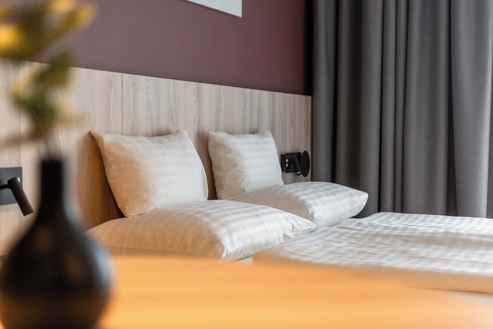 A hotel room with a wooden headboard, two beds with white bedding, a bedside table, and a vase with flowers.