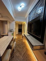 A modern hotel room with a herringbone wood floor, a flat-screen TV mounted on the wall, a dining area with a bench, and a small kitchenette.