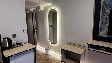 Ein modernes Hotelzimmer mit Fischgrätparkett, einer kleinen Küchenzeile, einem Spiegel mit integrierter Beleuchtung und einem großen gerahmten Kunstwerk an der Wand.
