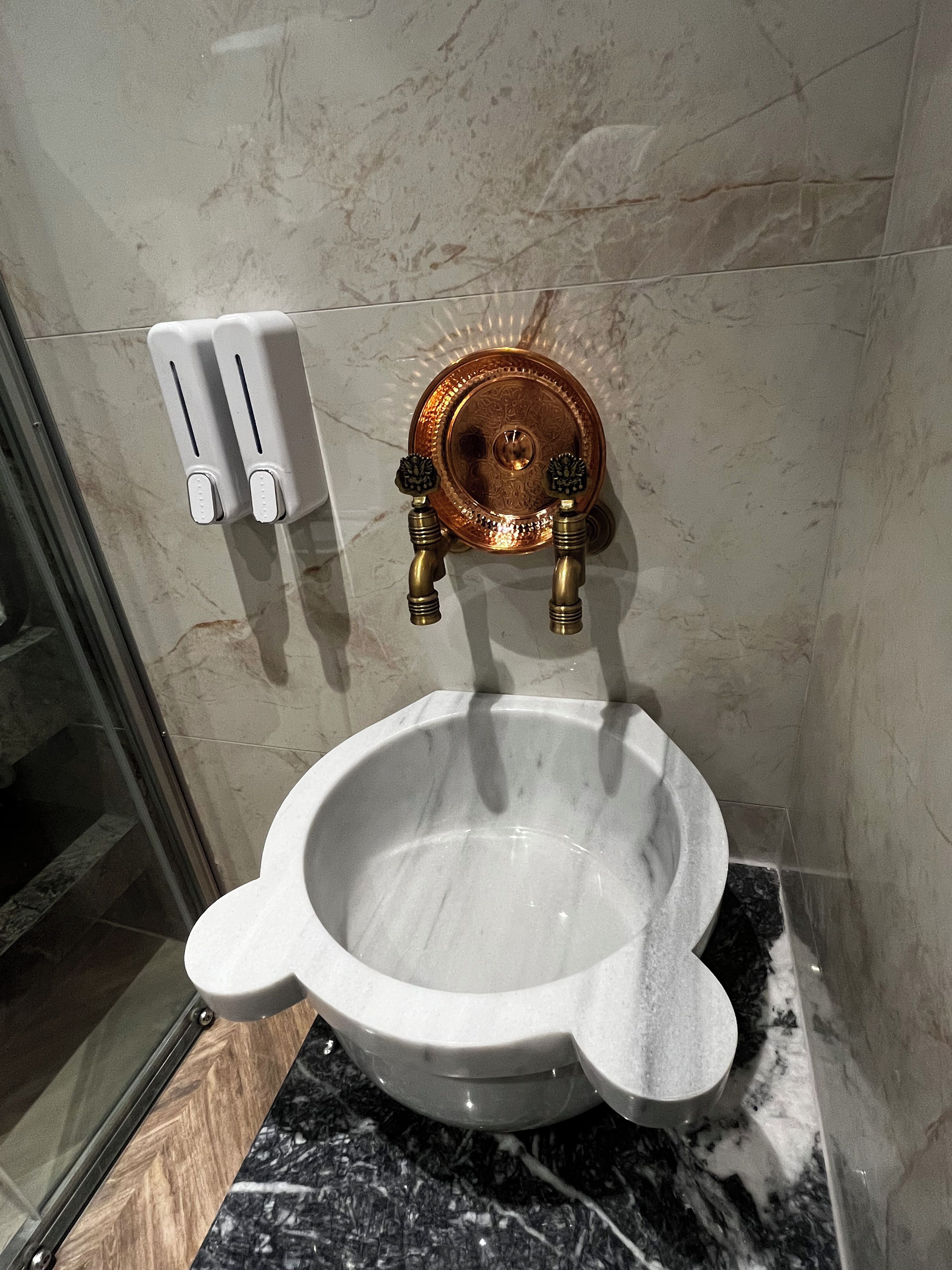 A bathroom with a white ceramic toilet, a copper-colored flush handle, and a soap dispenser on the wall.
