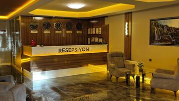 Uma área de recepção com balcão identificado como 'RESPEPSIYON', uma parede com relógios mostrando diferentes cidades, e um espaço de estar com poltronas e uma mesa de centro.