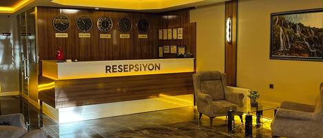 A reception area with a reception desk labeled 'RESPEPSIYON,' a wall with clocks showing different cities, and a seating area with armchairs and a coffee table.