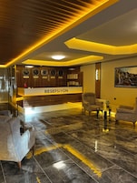 A reception area with a reception desk labeled 'RESPEPSIYON,' a wall with clocks showing different cities, and a seating area with armchairs and a coffee table.