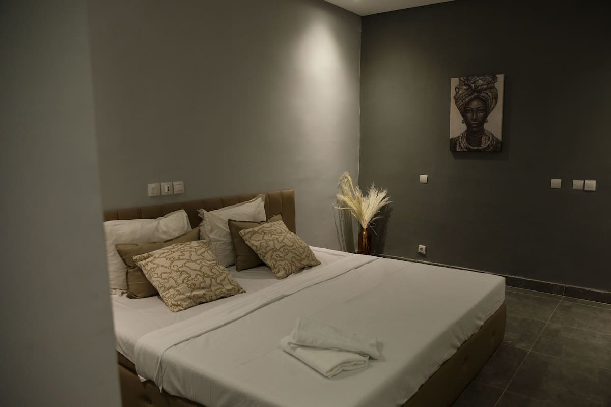 A neatly made bed with white and patterned pillows, a vase with dried plants, and a painting on the wall.