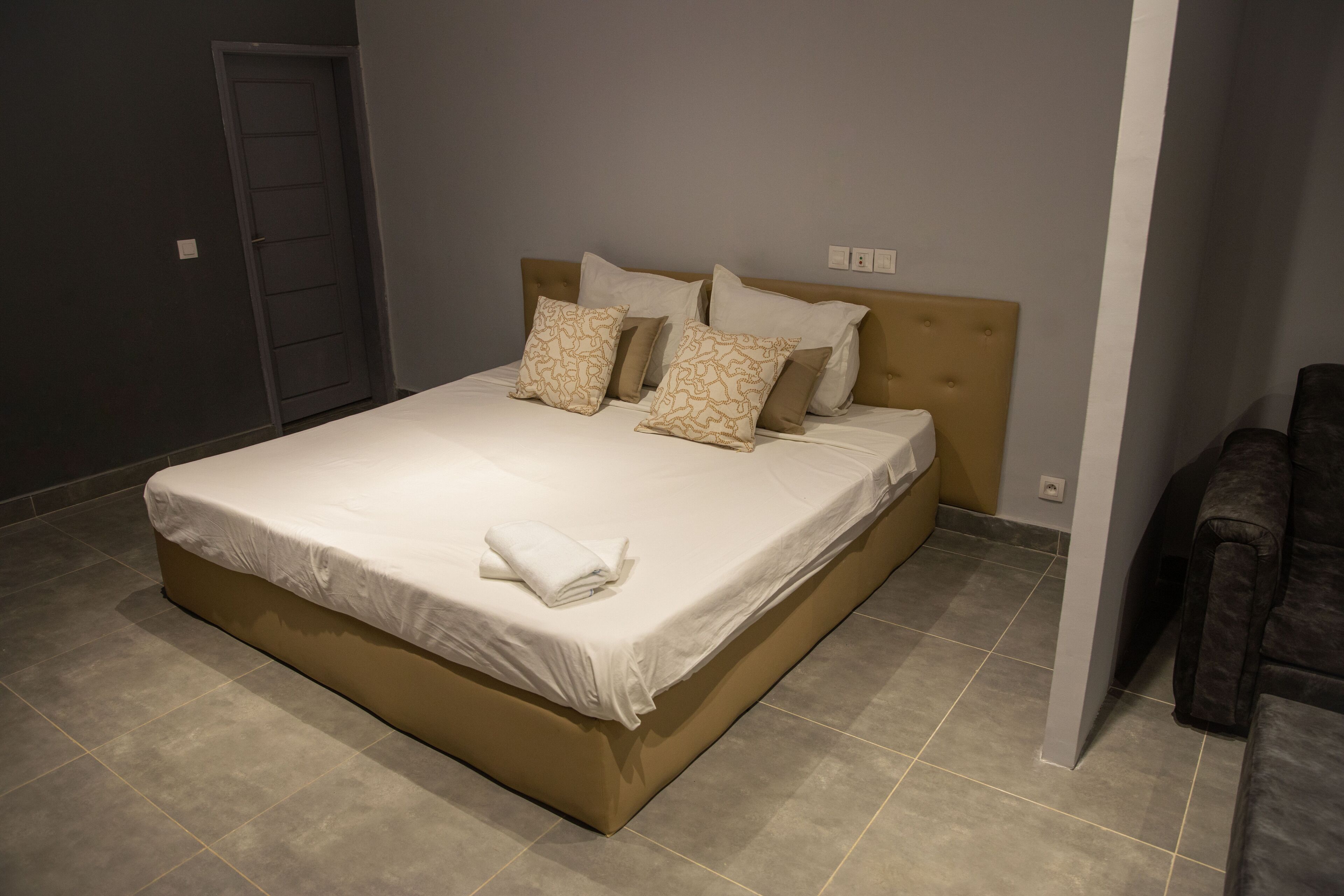 A neatly made bed with a beige headboard and white bedding, a dark door to the left, and a dark armchair to the right.