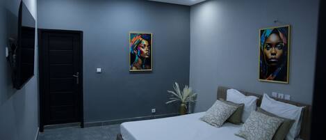 A modern bedroom with a large bed, two wall-mounted paintings, a potted plant, and a flat-screen TV.