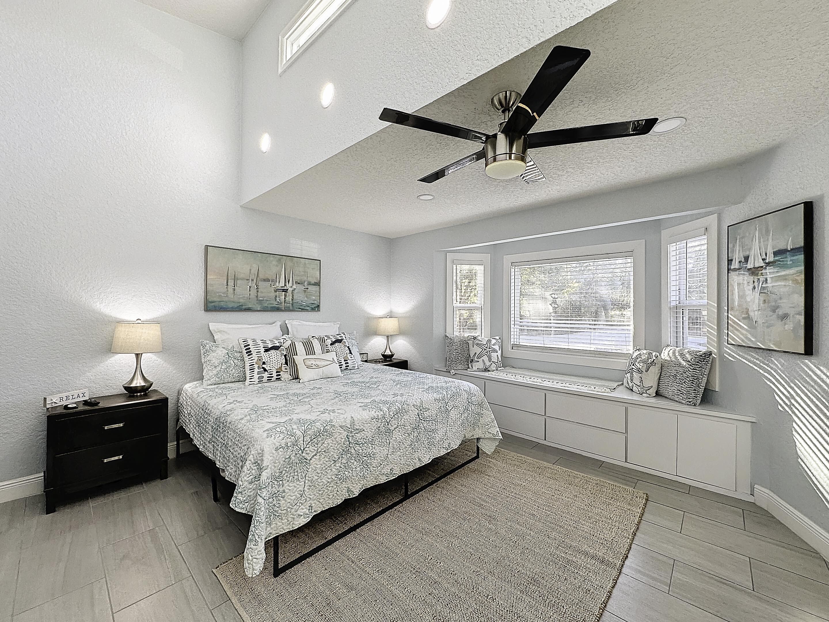 Une chambre à coucher avec un lit, des tables de chevet, un ventilateur de plafond et des œuvres d’art accrochées aux murs.