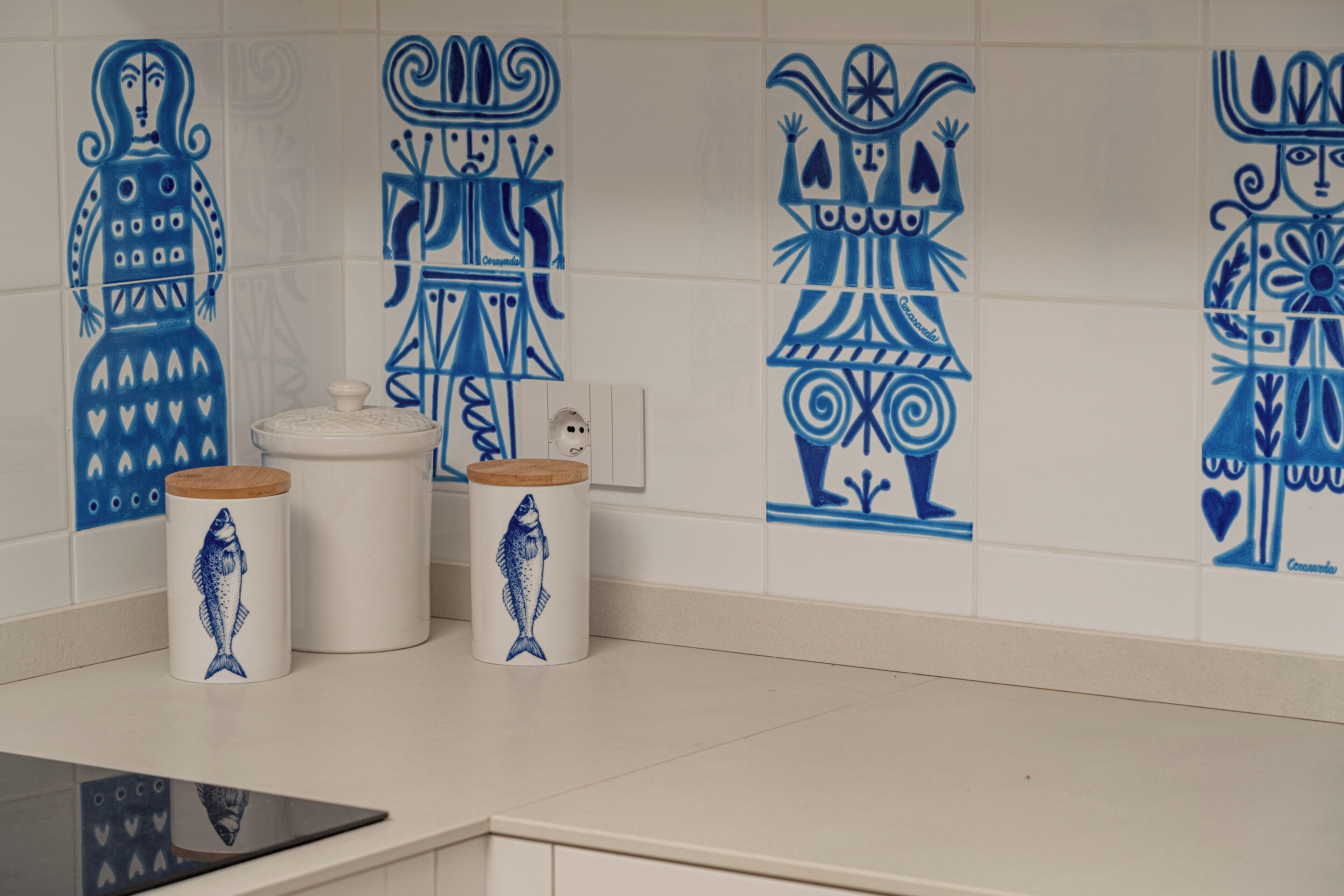 A kitchen with white tiles and blue decorative patterns. There are two white containers with wooden lids and fish designs.