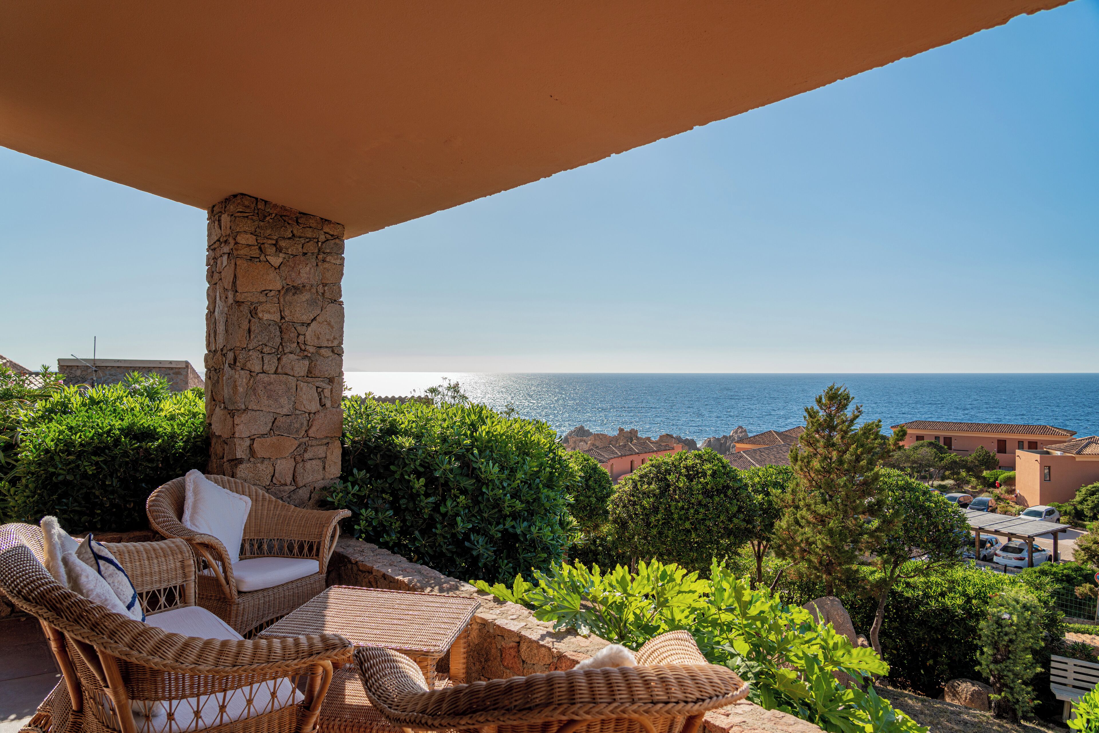 A balcony with wicker furniture, a stone pillar, and a view of the sea and buildings.