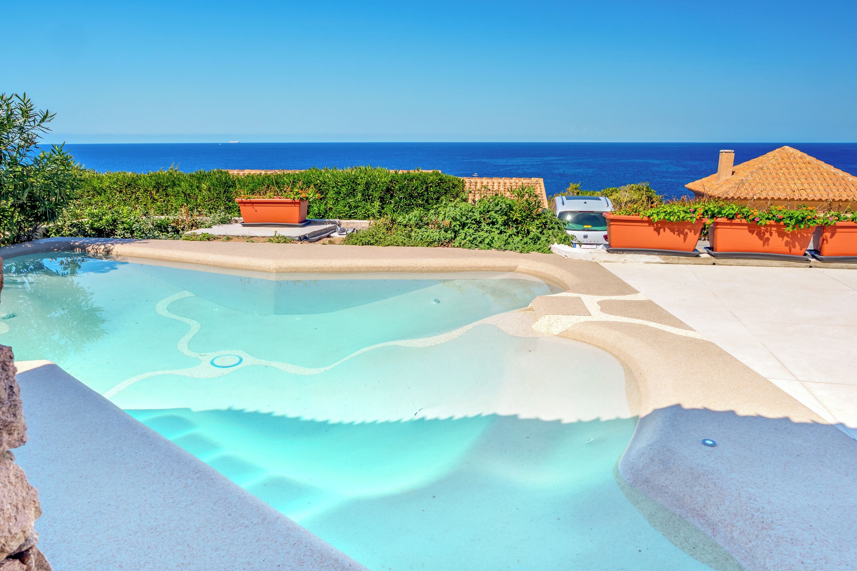 A pool with a clear blue edge, surrounded by potted plants and a view of the ocean.