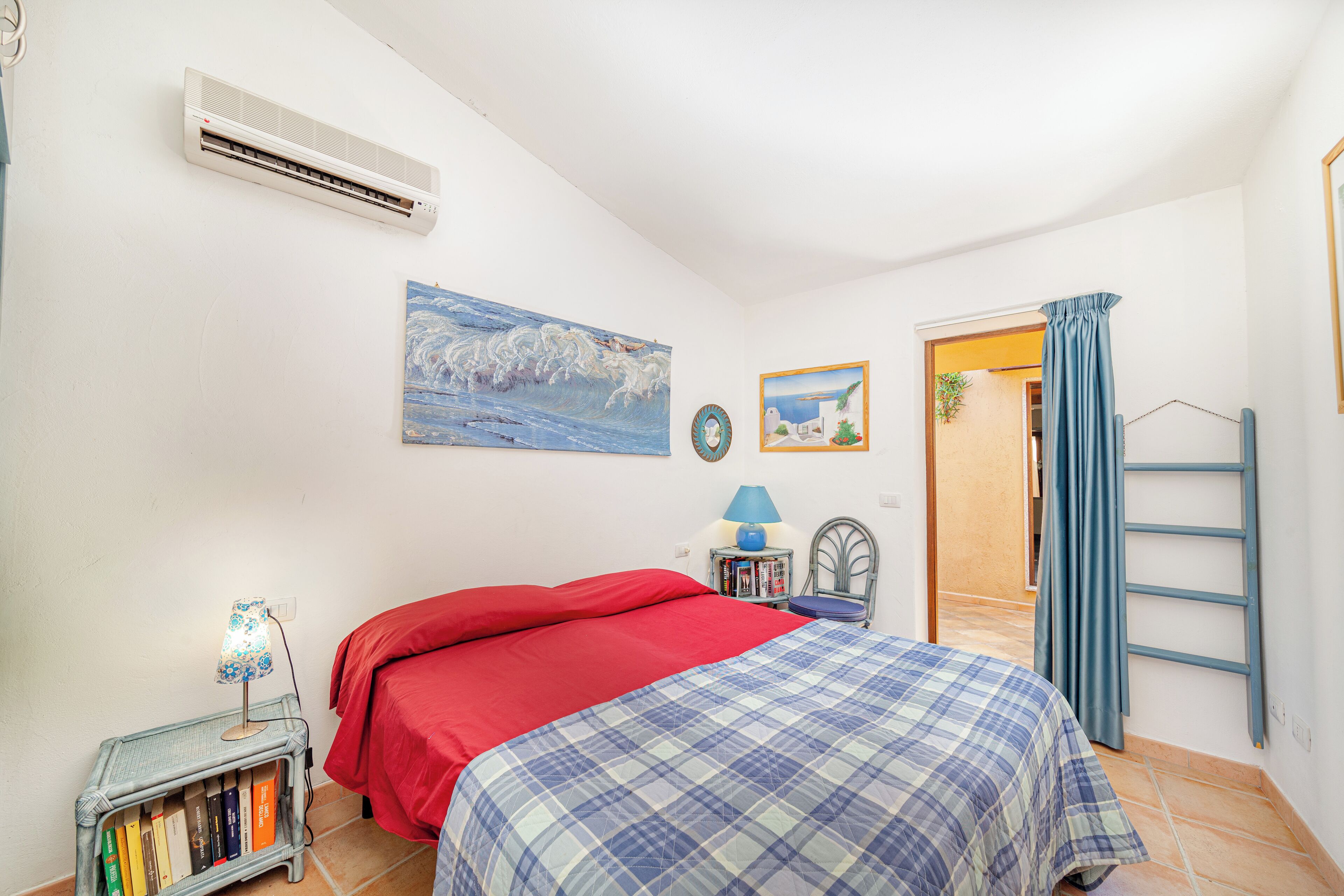 A bedroom with a bed, a nightstand with books, a lamp, a painting, and an air conditioning unit.